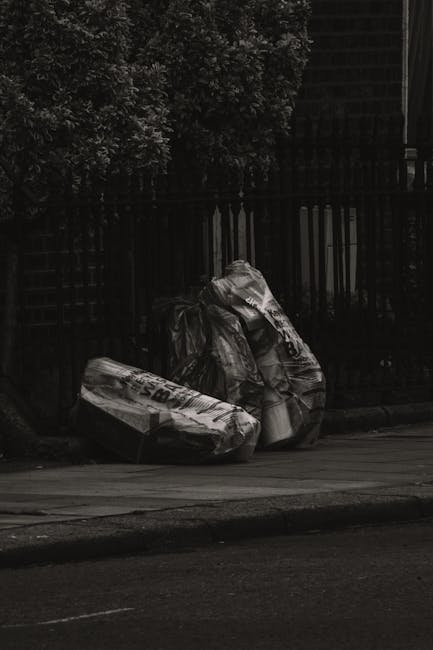A pair of large, black garbage bags filled with waste material, placed on a sidewalk next to a curb. The bags are made of thick plastic and appear to be tightly packed, with some creases and folds visible on their surfaces. They are positioned in front of a black metal fence with vertical bars, which runs along a residential or urban area. Behind the fence, there are dense leafy trees with dark foliage, indicating a shaded environment. The ground is paved with asphalt, and the scene appears to be outdoors during daylight, though the image is in black and white, emphasizing the textures and contrasts between the dark bags, fence, and surrounding environment. This scene illustrates typical private waste collection, suitable for services like those offered by Rubbish Removal Brompton, and highlights the importance of proper disposal for effective rubbish removal in urban areas.