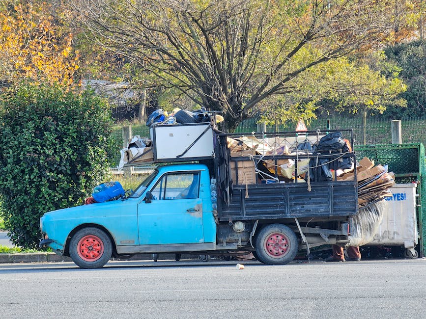 A blue flatbed truck parked alongside a roadway, loaded with a large assortment of waste and recyclable materials, including cardboard boxes, plastic bags, and various household items. The truck's surface shows signs of wear with visible dirt and minor rust, and it has red-painted wheels. The loading area is surrounded by metal railings, partially supporting the stacked debris. In the background, a tree with autumn foliage and a green hedge are visible, indicating an outdoor, residential area. The scene appears to depict a private rubbish removal effort, characteristic of independent waste collection services, with the vehicle positioned on a paved street next to a sidewalk and grass verge. The overall atmosphere is calm, and the natural lighting highlights the textures of the materials and the details of the truck's surface.