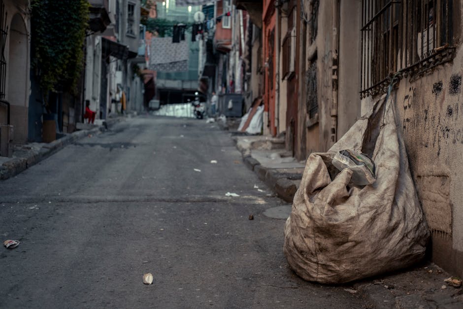 A worker dressed in high-visibility orange and reflective safety gear, wearing an orange cap, is seen from above cleaning a sidewalk area. The person is holding a broom and appears to be sweeping a collection of mixed waste materials, which includes dried leaves, crumpled paper, a white disposable cup, and other miscellaneous litter. The waste is gathered along the edge of a paved path, adjacent to a rough concrete curb that separates the walkway from an asphalt road surface. The scene is outdoors, with natural daylight illuminating the area, and the worker’s attire suggests a professional rubbish removal activity. The presence of collected debris and the worker’s focus on clearing it indicate an ongoing task of waste collection or street hygiene maintenance, aligning with private rubbish handling services such as those provided by Rubbish Removal Brompton to maintain cleanliness in urban environments.